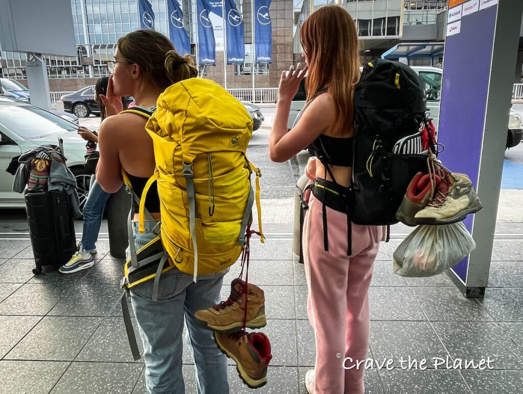 2 girls with hiking backpacks at frankfurt airport one with yellow bag and both with hiking boots hanging off
